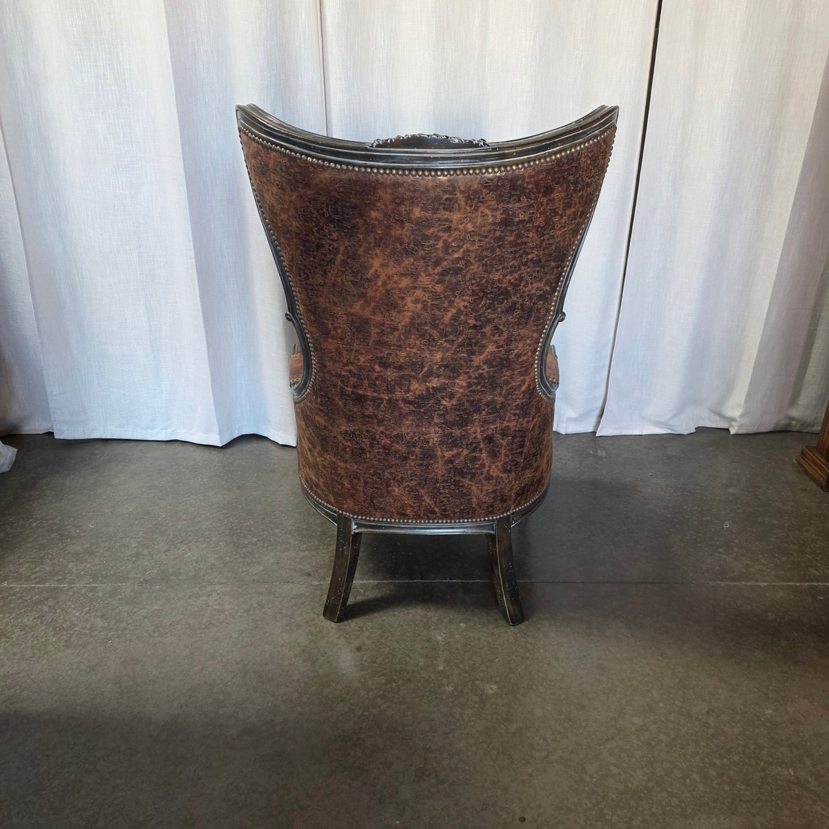 A tall, curved-back wingback armchair upholstered in distressed brown leather, shown from the back against a white curtain backdrop on a concrete floor.