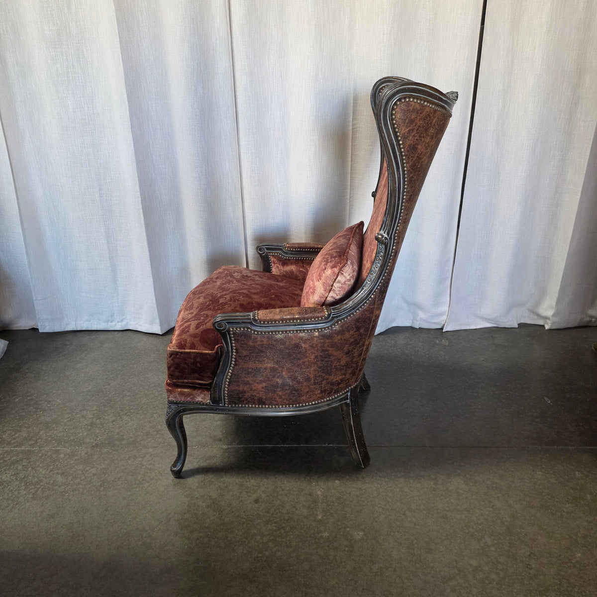 A vintage-style wingback armchair upholstered in distressed reddish-brown velvet with a dark carved wooden frame and nailhead trim, shown against a white curtain backdrop.