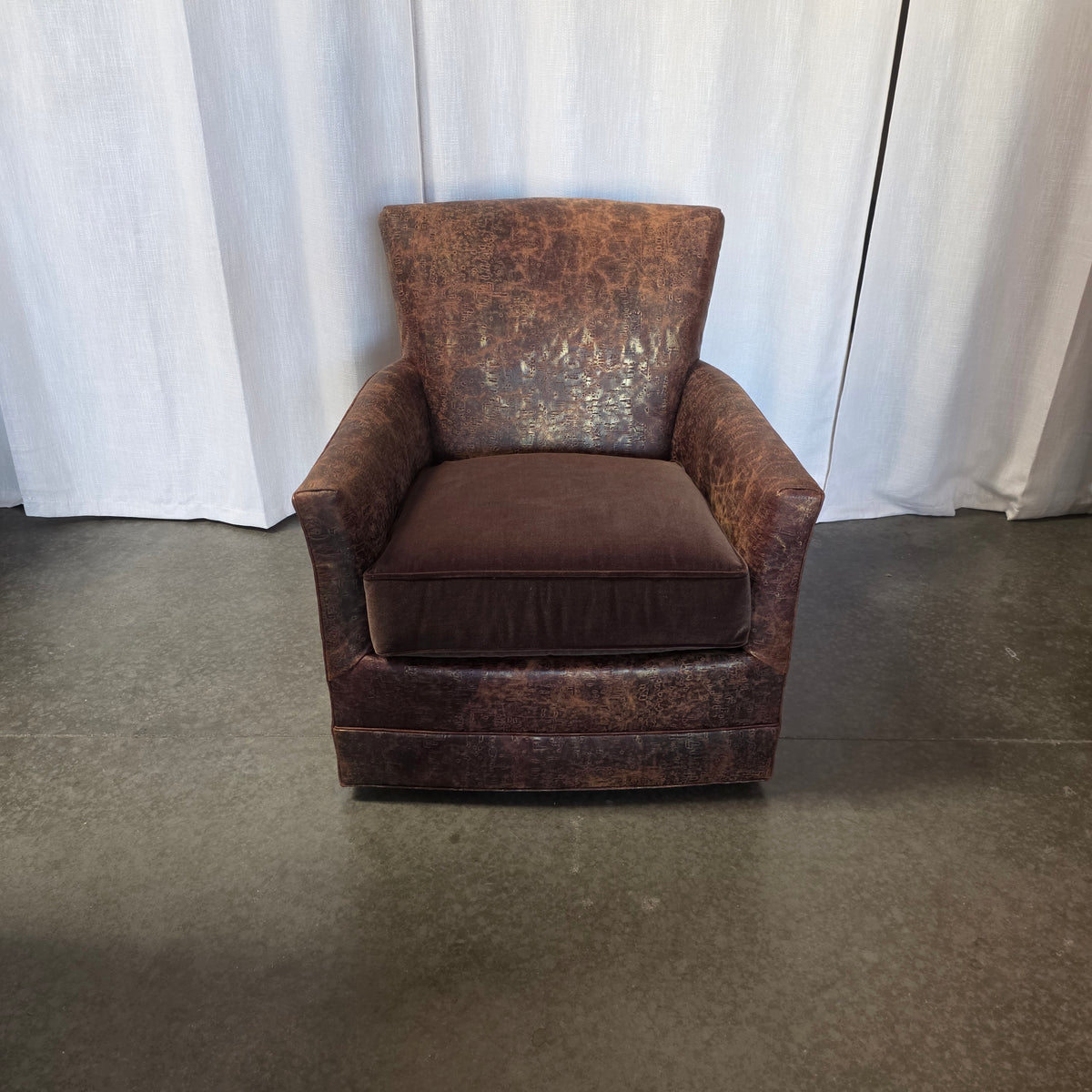 A distressed brown leather armchair with a matching brown cushion, positioned in front of a white curtain backdrop on a gray concrete floor.