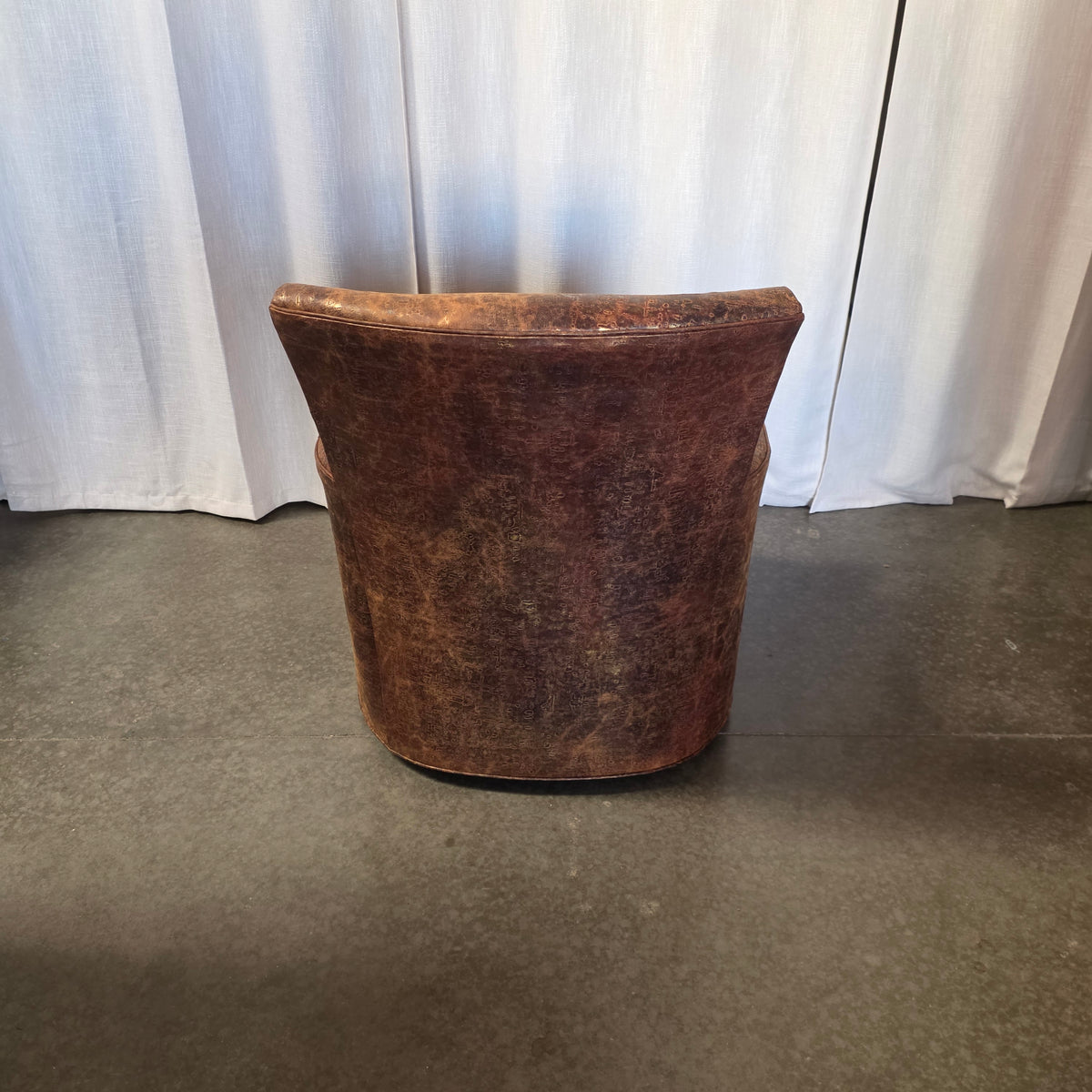 A rustic, distressed brown leather bucket chair or stool with a curved back, shown from the rear against a backdrop of white curtains and a gray concrete floor.