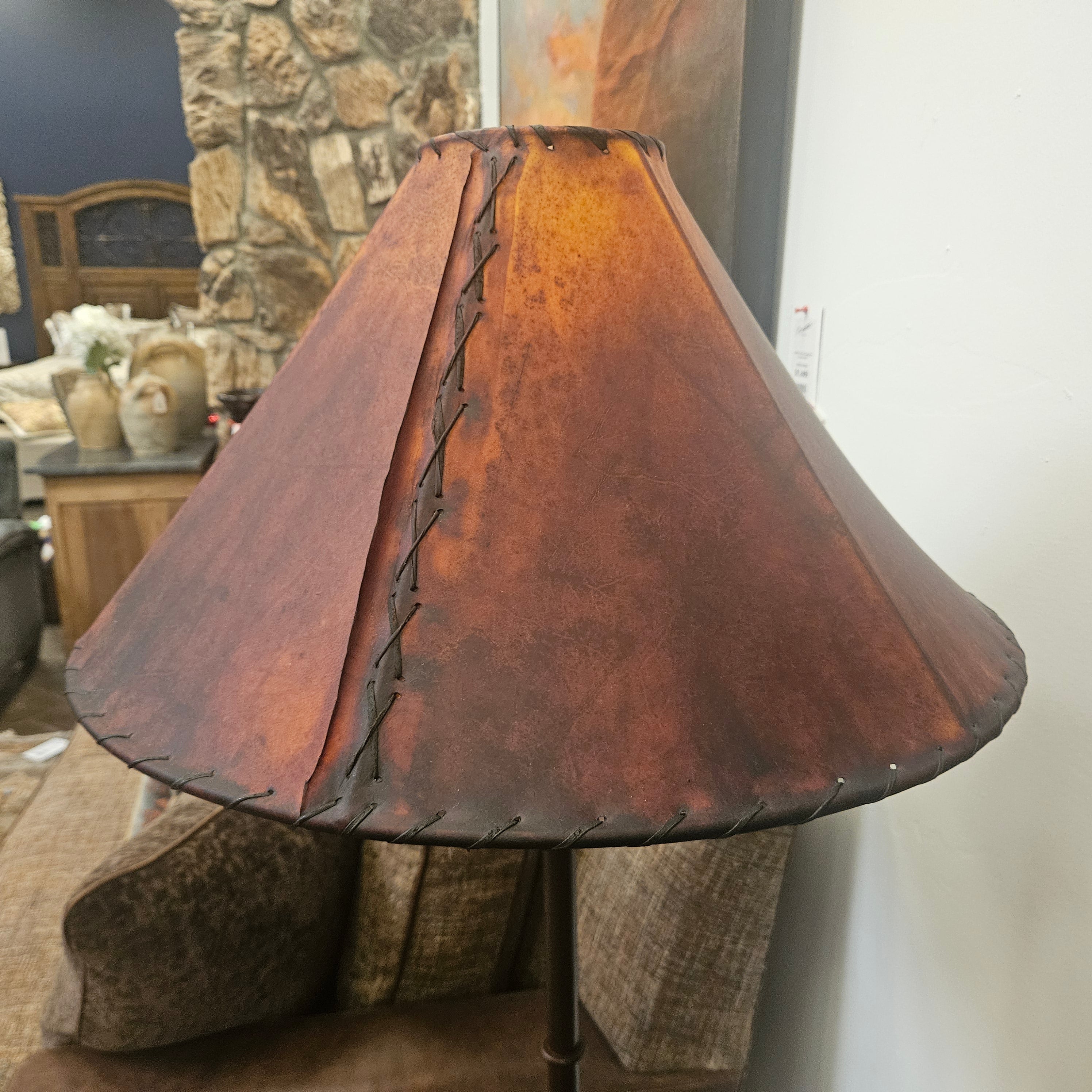 A rustic brown leather lampshade with dark stitched detailing along the seams, displayed on a table in a home decor store setting.