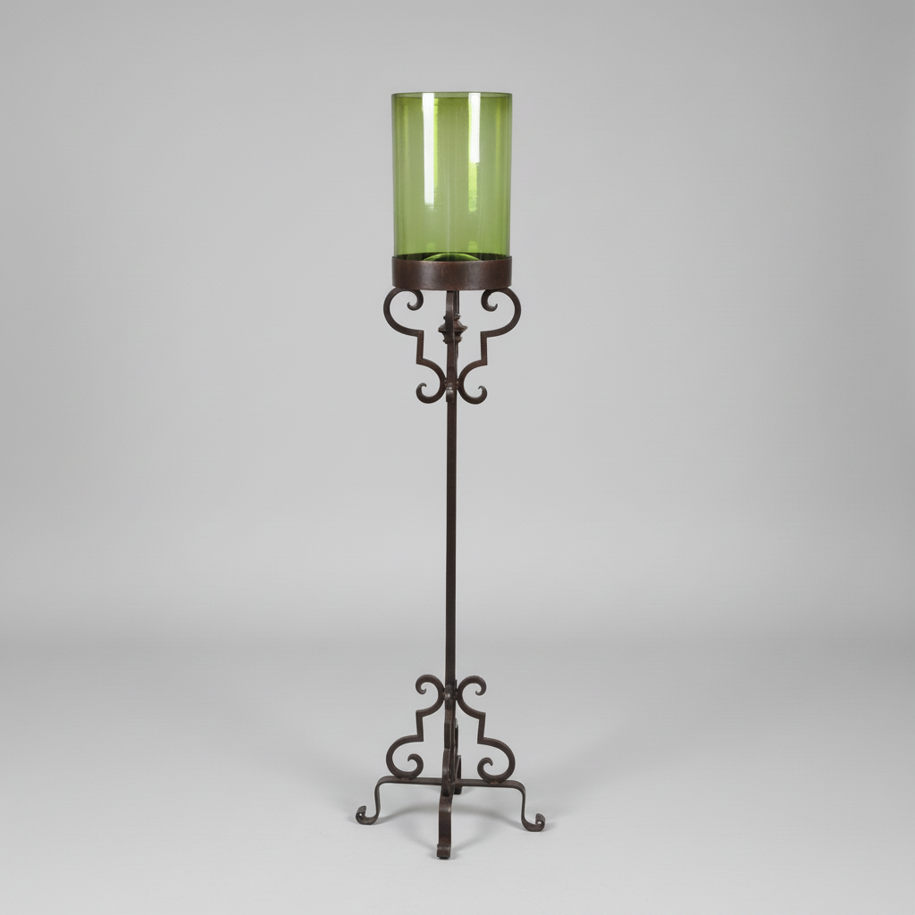 A tall green glass candle holder with a dark metal scrollwork stand, photographed against a neutral light gray background.