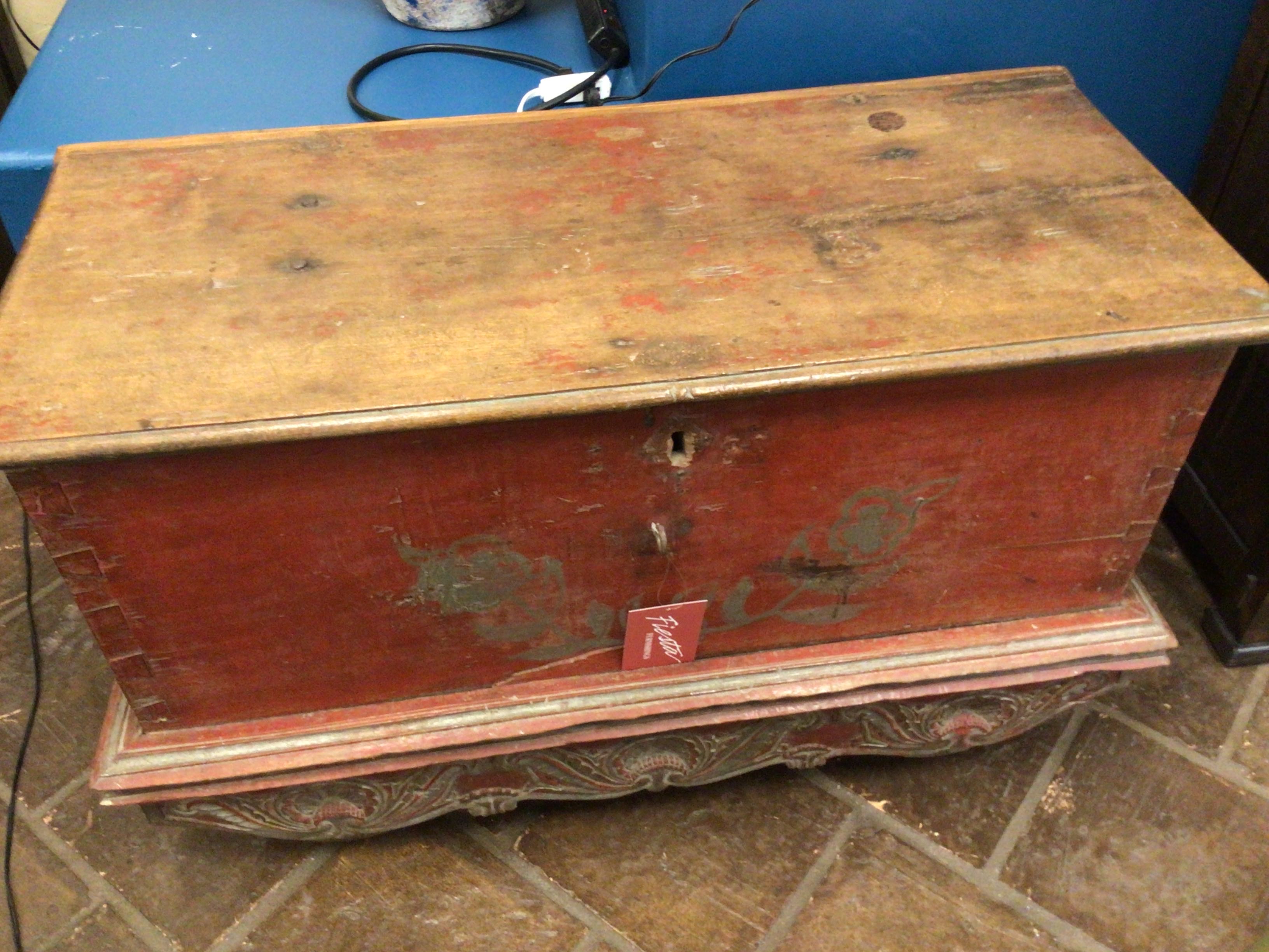 Old wooden chest with red paint on a tiled floor against a blue wall.