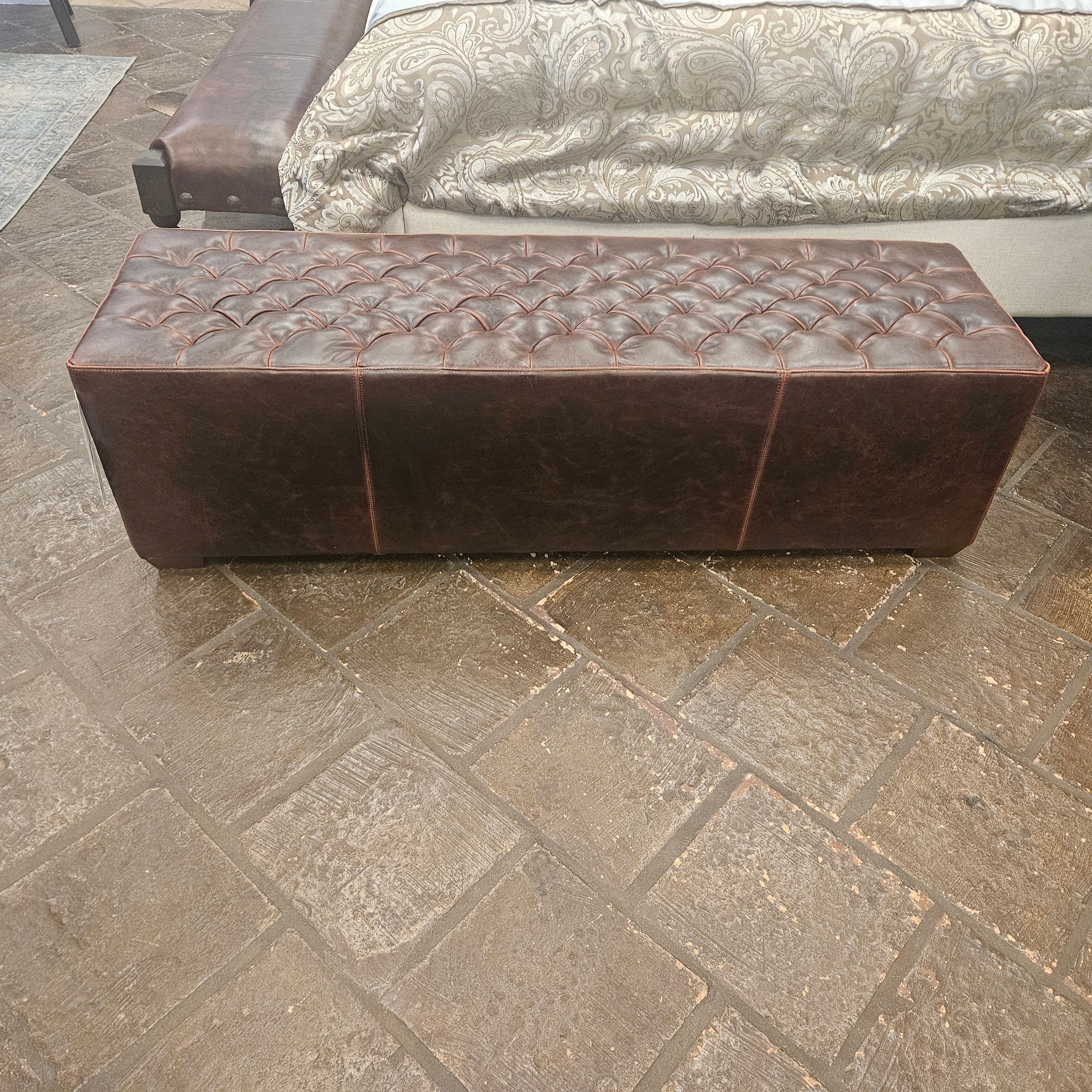 Brown leather ottoman on a stone floor with patterned bedding in the background