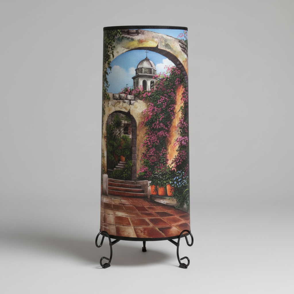 A cylindrical table lamp with a printed scenic design of a Mediterranean-style courtyard with arches, terracotta tiles, and pink flowers, displayed on a black metal stand against a neutral gray background.
