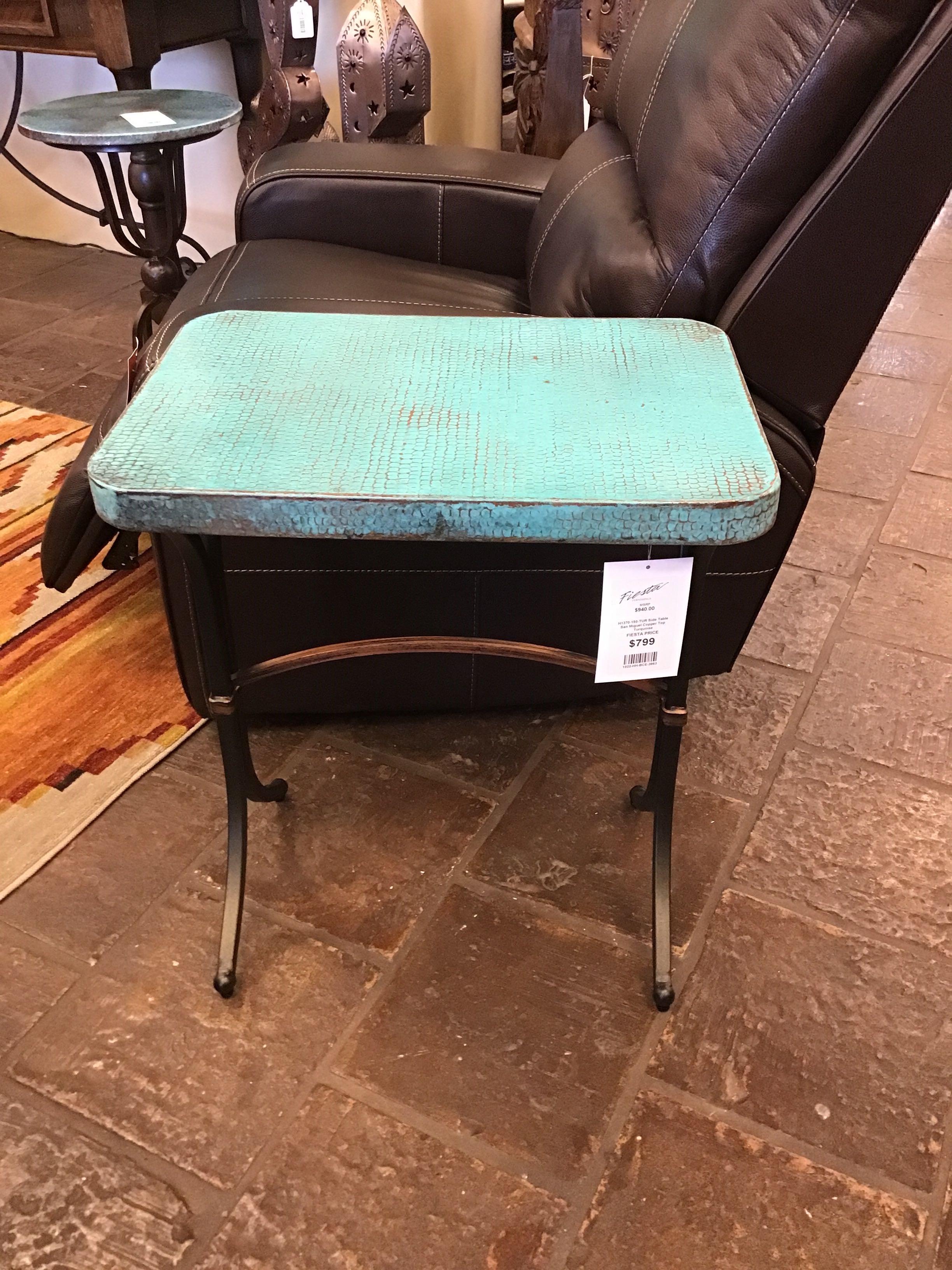 Turquoise ottoman with a label in front of a black leather chair on a stone floor.