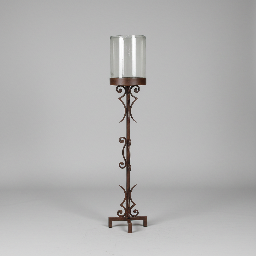 A dark brown wrought iron candle holder with an ornate scrollwork design, featuring a clear glass cylinder shade on a neutral gray background.