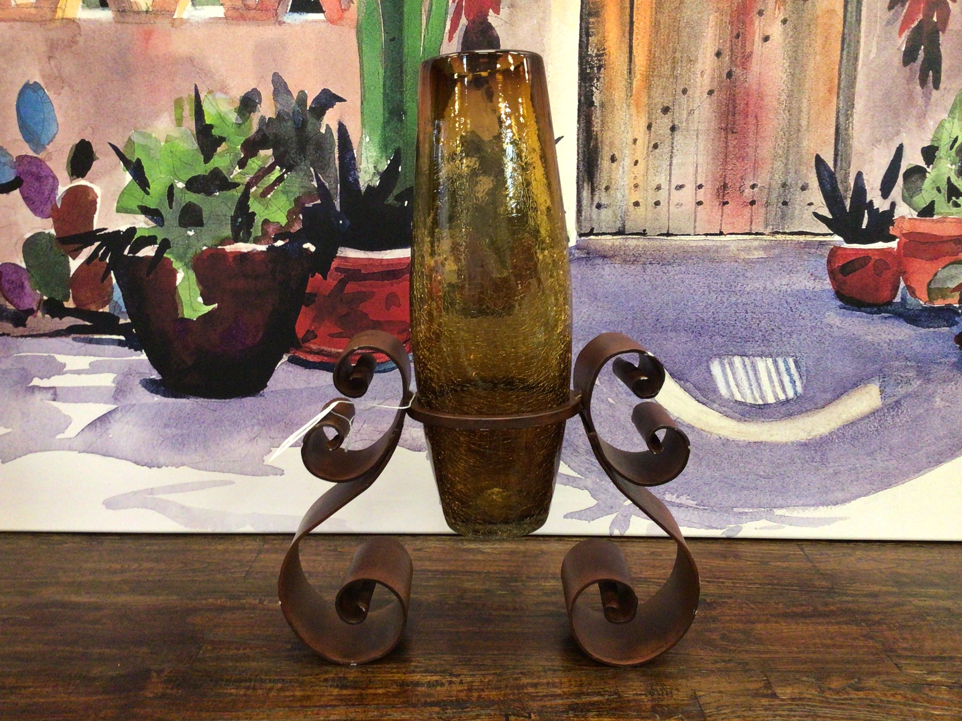 Decorative metal stand with a textured vase on a wooden surface, with a painting of plants in the background.