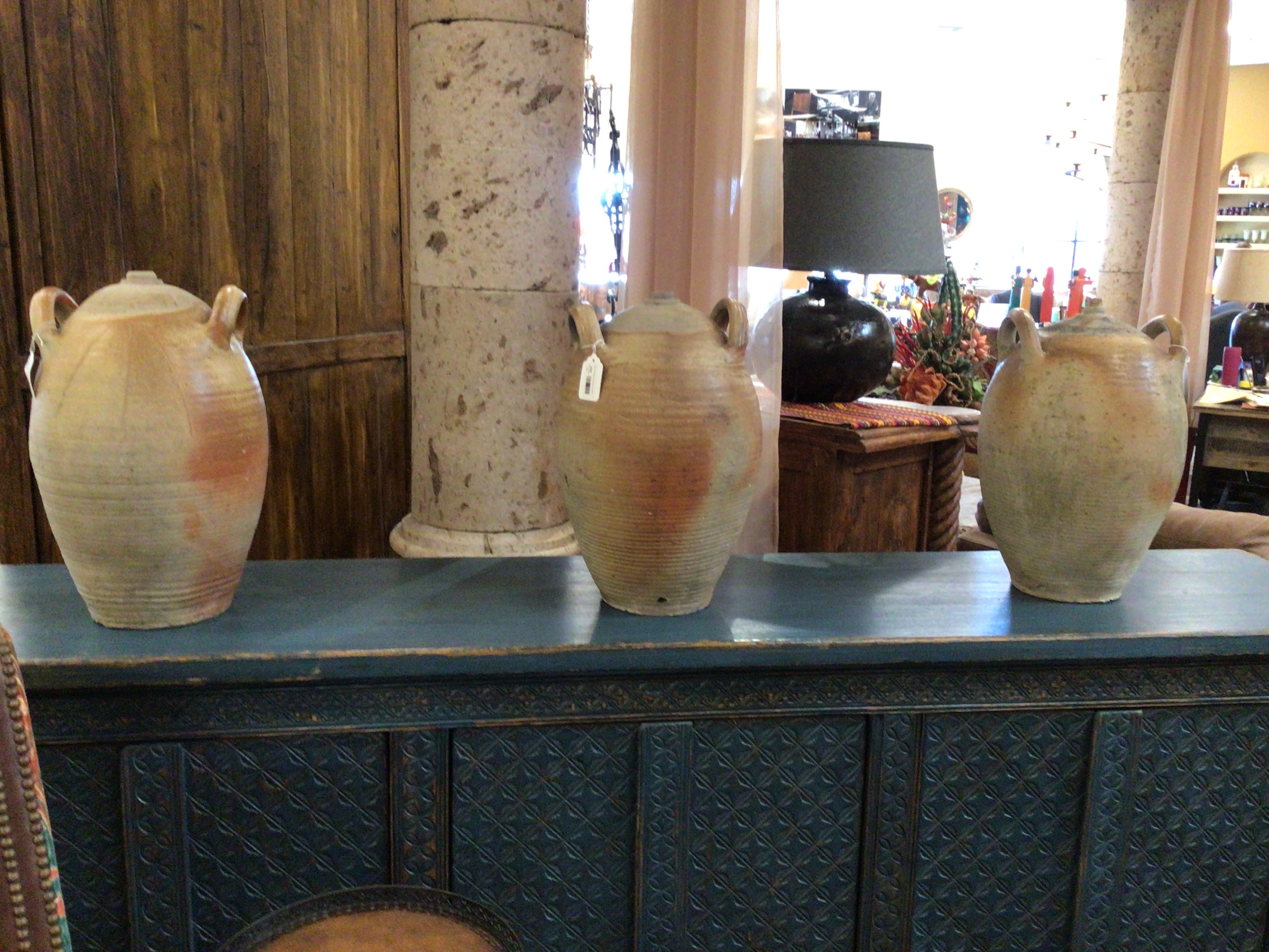 Three ceramic jars on a blue surface with a wooden cabinet and decorative items in the background.