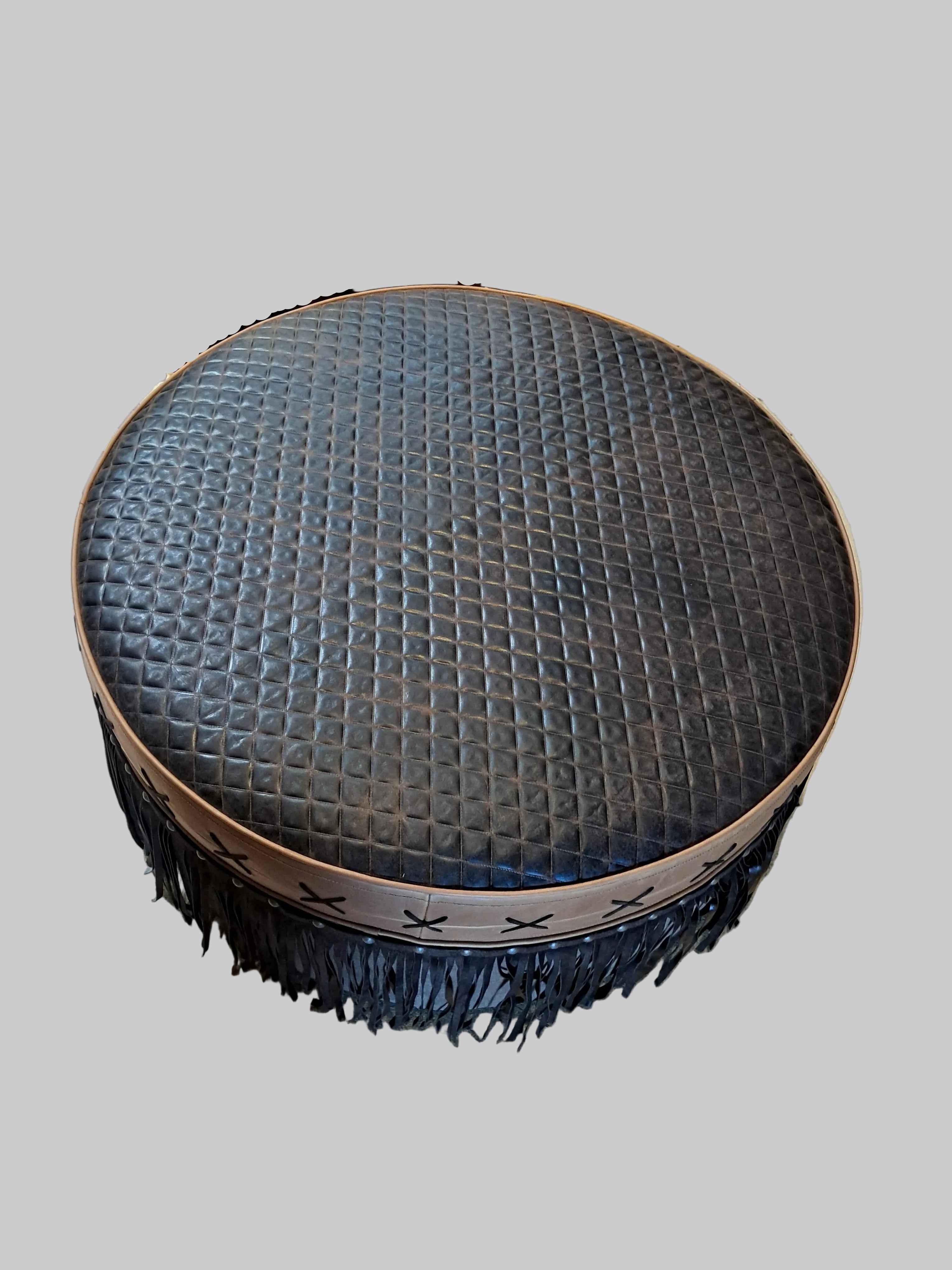 Round ottoman with textured top and fringe trim on a concrete floor.