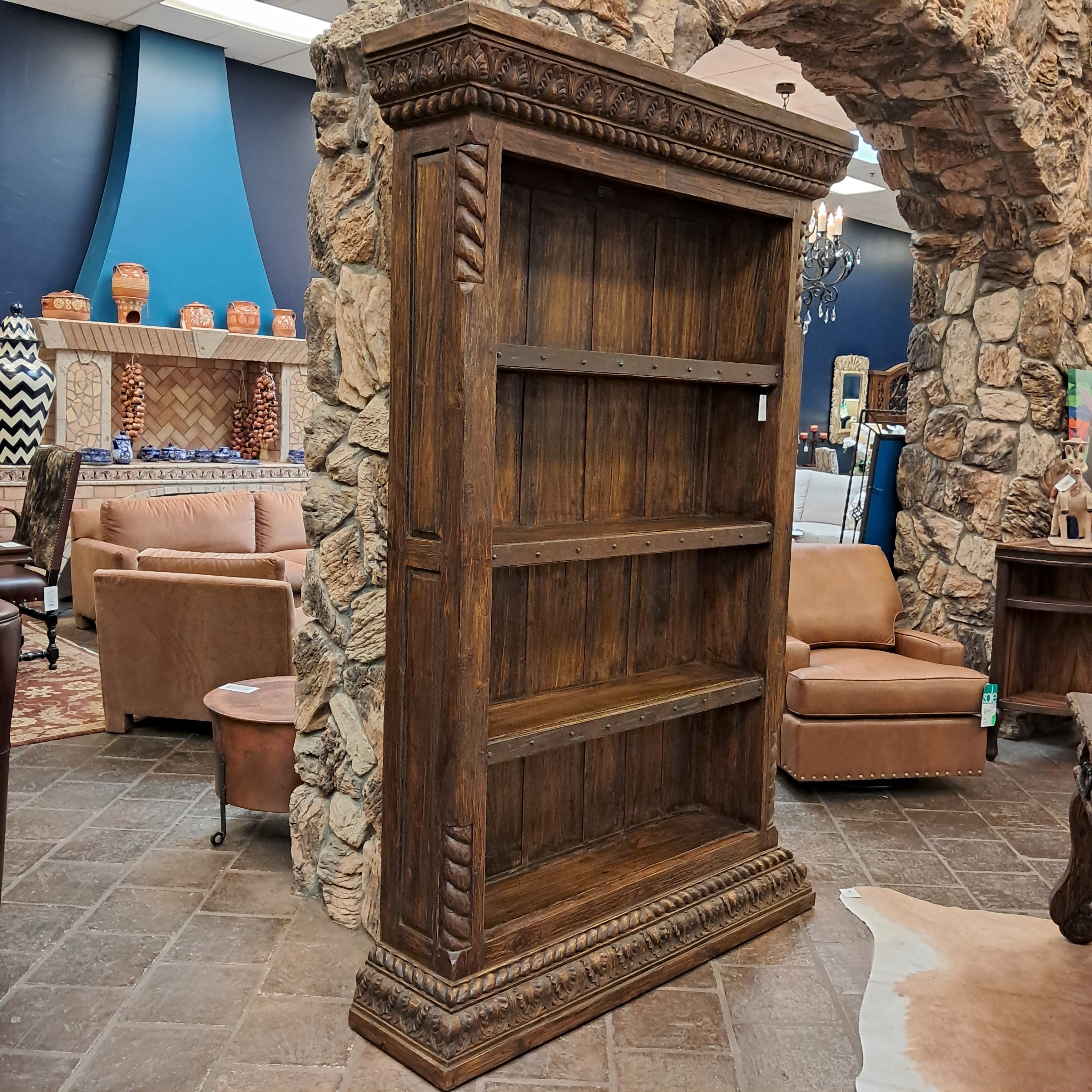 A tall, rustic dark brown wooden bookcase with carved details and three shelves, positioned in a rustic showroom with stone arches and leather furniture in the background.