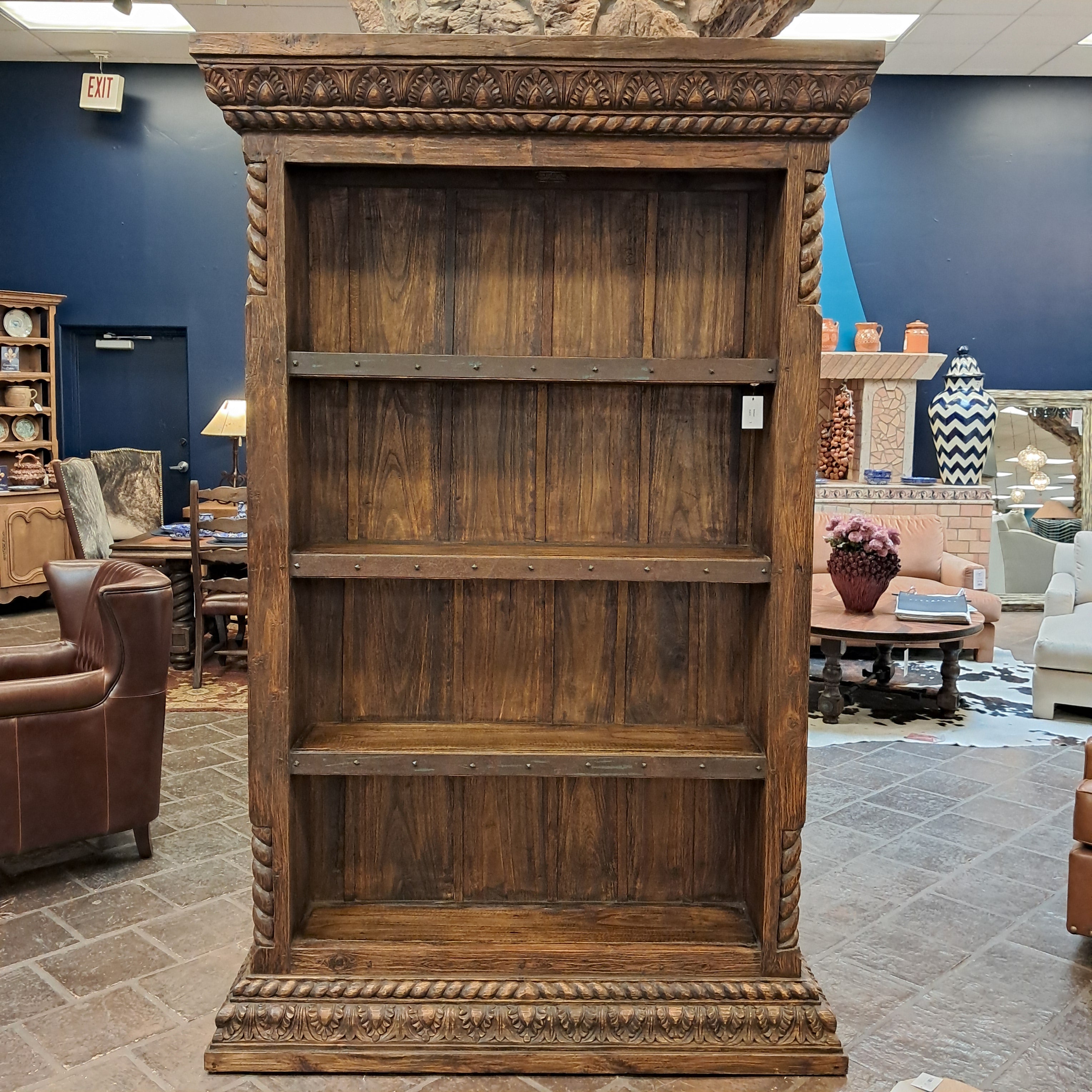 A large rustic wooden bookcase with three open shelves, ornate carved side panels, and metal bracket hardware, photographed in a furniture showroom.