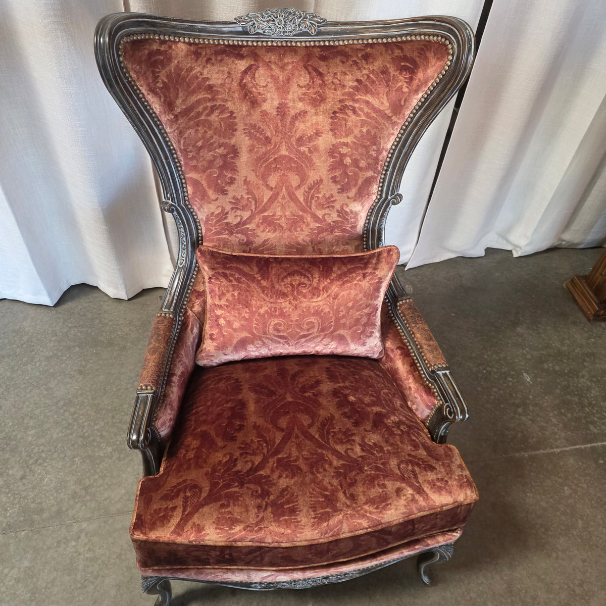 An antique-style wingback armchair with a matching lumbar pillow, upholstered in a dusty rose damask velvet fabric with a dark carved wooden frame, set against a backdrop of white curtains and a concrete floor.