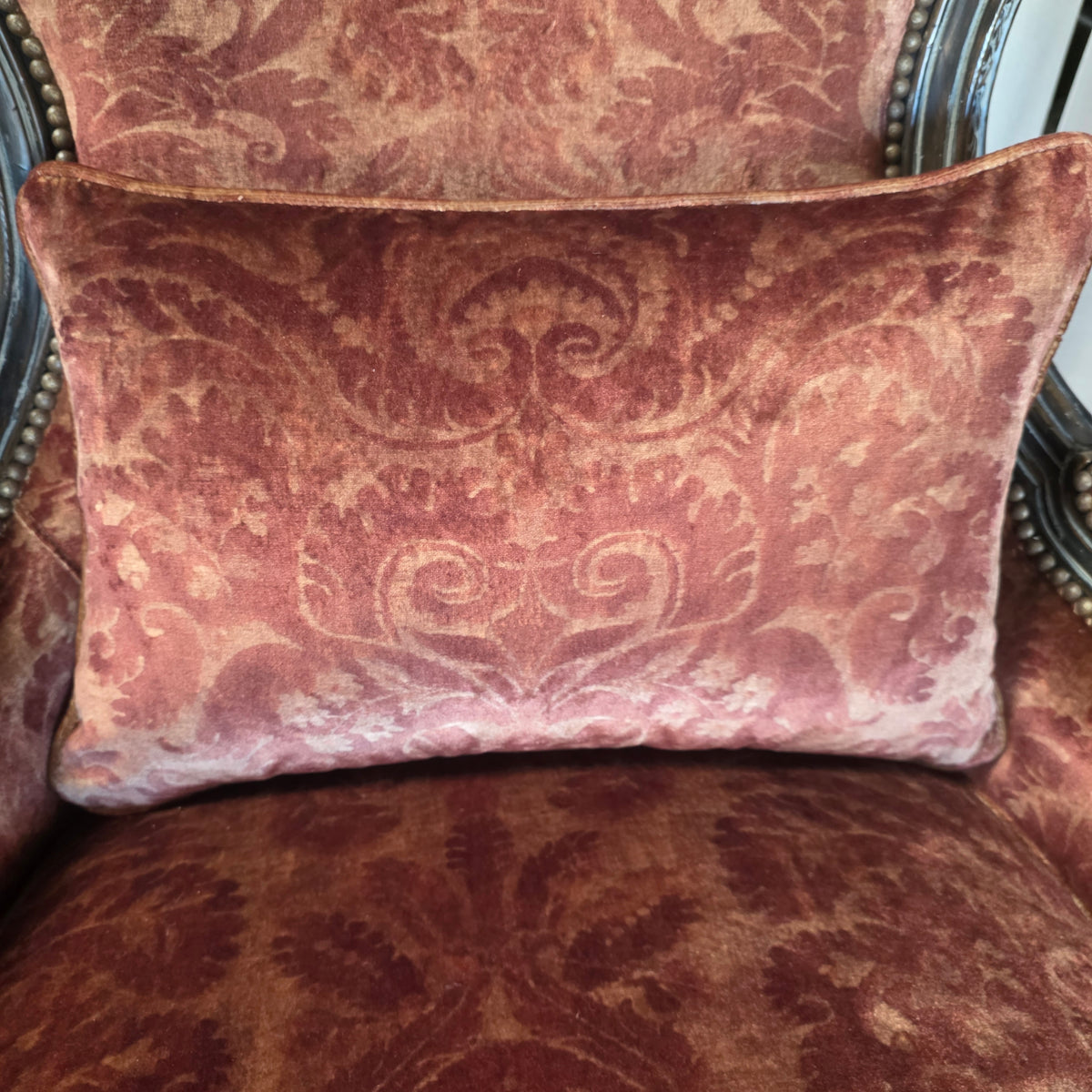 A rectangular lumbar throw pillow with a dusty rose damask pattern, resting on an upholstered armchair with matching fabric.
