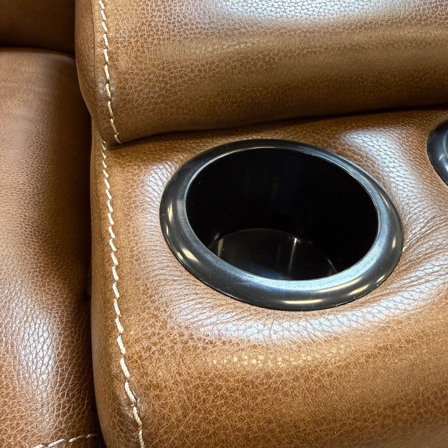 Close-up of a brown pebbled leather recliner chair with a circular black cup holder insert and white contrast stitching.