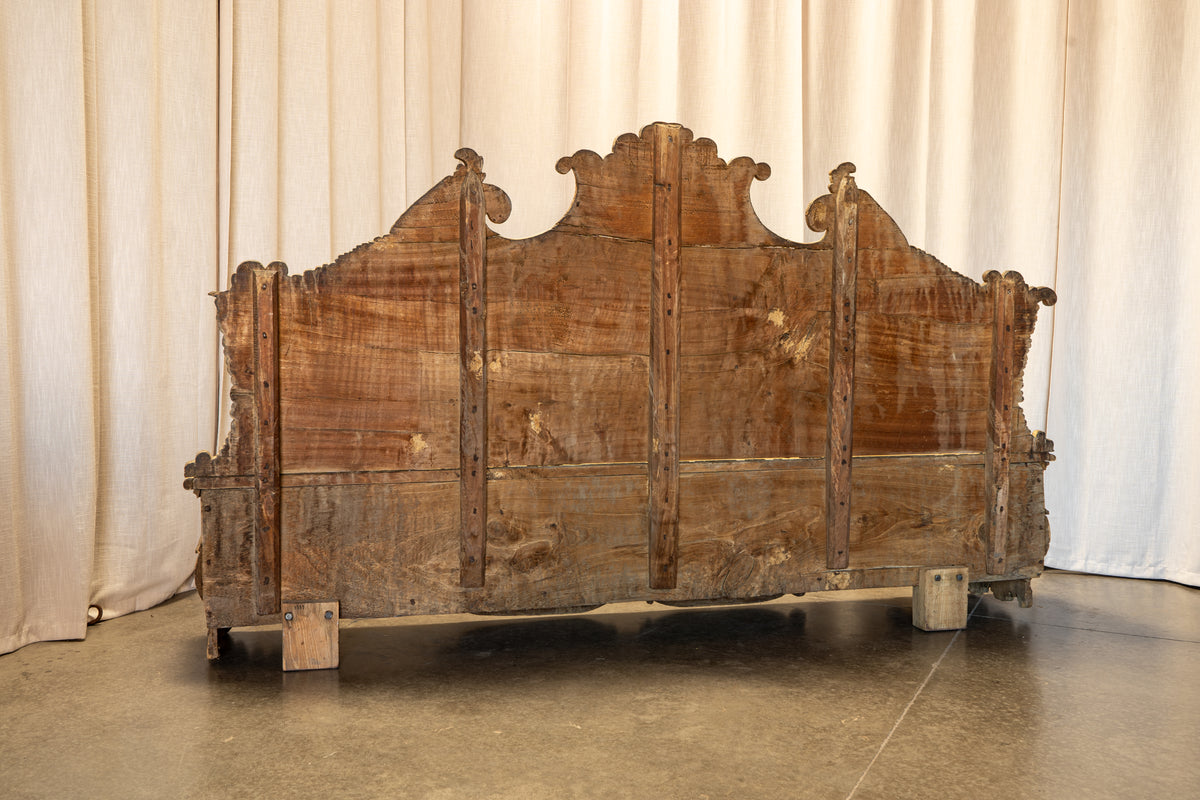A large, rustic wooden headboard with a distressed finish, featuring a high arched crest and carved details, photographed against a neutral cream curtain backdrop.