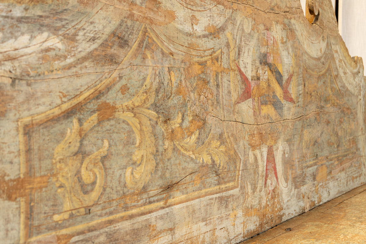 Close-up of an aged painted wooden panel or wall decoration with faded gold, beige, gray, and red decorative motifs and visible cracks and wear.