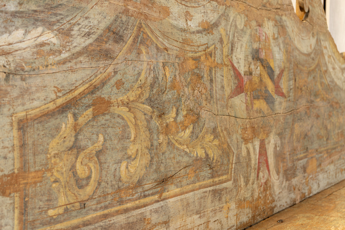 Close-up of an aged painted wooden panel or furniture surface with faded scrollwork and decorative motifs in beige, gray, brown, and small red accents.