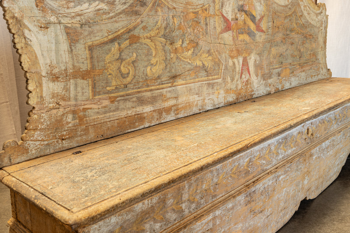 An antique painted wooden chest or bench with a large decorative backrest panel, showing weathered beige, gold, and faded red painted details.