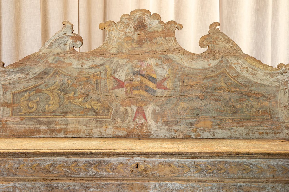 Close-up of an ornate antique wooden headboard or bench backrest with a distressed painted finish, featuring a central heraldic-style motif in muted red, gold, and gray tones against a cream curtain background.
