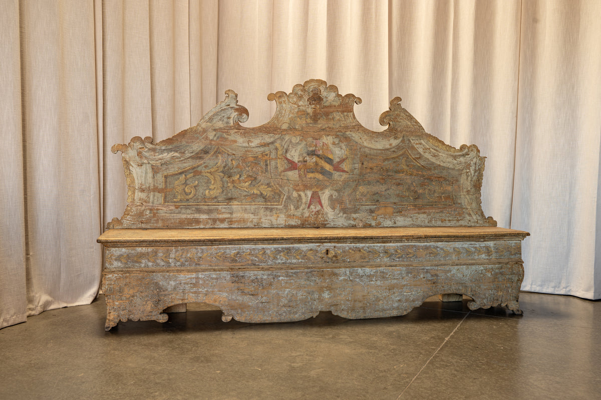 An antique carved wooden bench or bed frame with a highly decorative, distressed headboard featuring faded blue, green, and gold painted details, set against a neutral draped curtain backdrop.