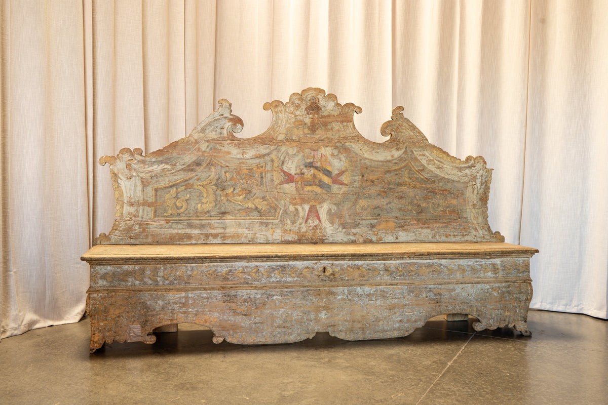 An antique carved wooden bench with a highly detailed, painted crest backrest, set against a neutral draped fabric backdrop.