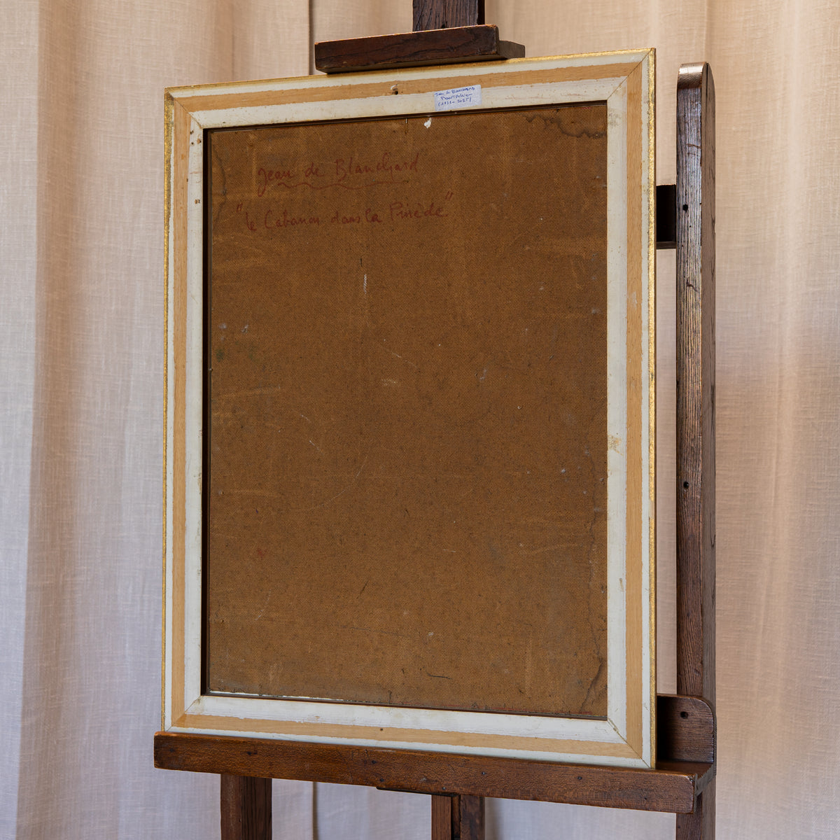 The back of a framed artwork on a wooden easel, showing a brown backing board with faint handwritten text in the upper left and a distressed off-white and gold frame against a beige curtain background.
