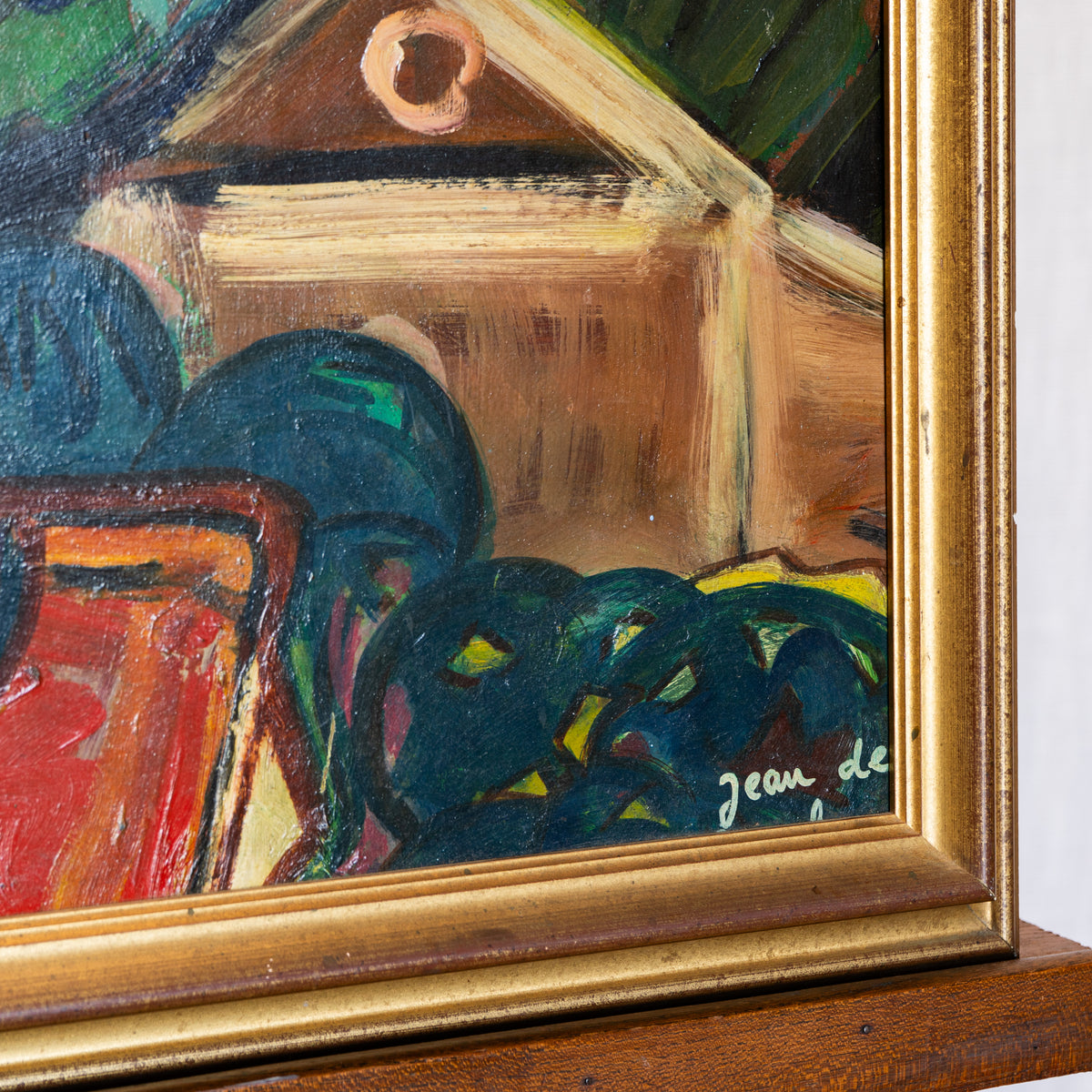 Close-up of a framed painting showing an abstract still life with dark blue-green forms, a tan box, red and orange shapes, and a gold frame. A signature reading 'Jean de' is visible near the lower right corner.