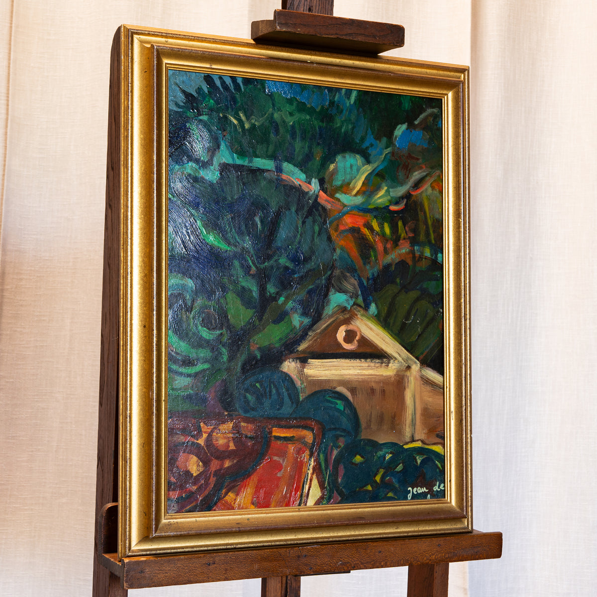 A framed oil painting depicting a rustic house surrounded by dark green trees and foliage, displayed on a wooden easel against a light beige curtain background.