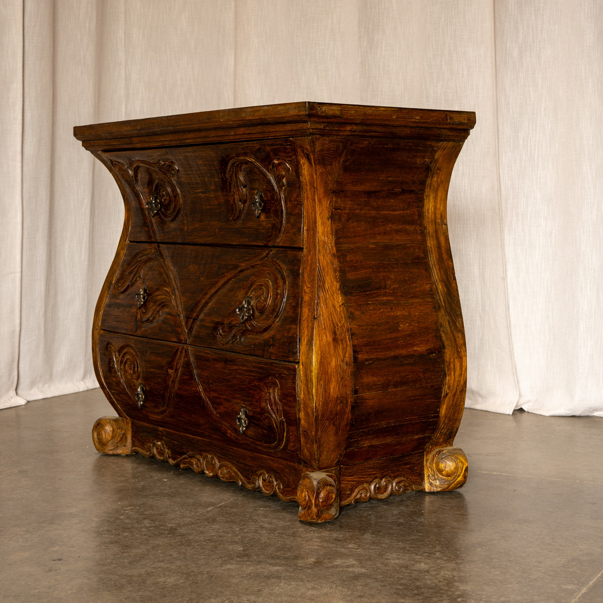 An ornate dark brown wooden chest of drawers with carved floral details and curved legs, photographed against a draped white fabric backdrop. Jason Scott Furniture - Angelina Dresser -Fiesta Furnishings. 