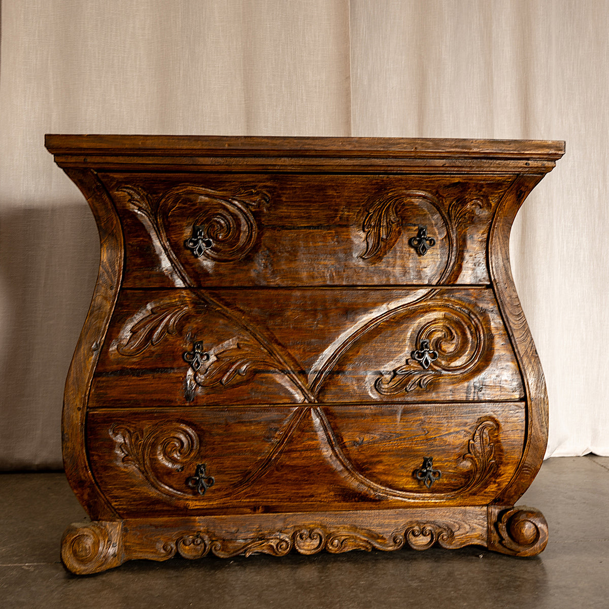 An antique carved wooden chest of drawers with ornate scrollwork and dark metal knobs, set against a neutral beige curtain backdrop. Jason Scott Furniture - Angelina Dresser -Fiesta Furnishings. 