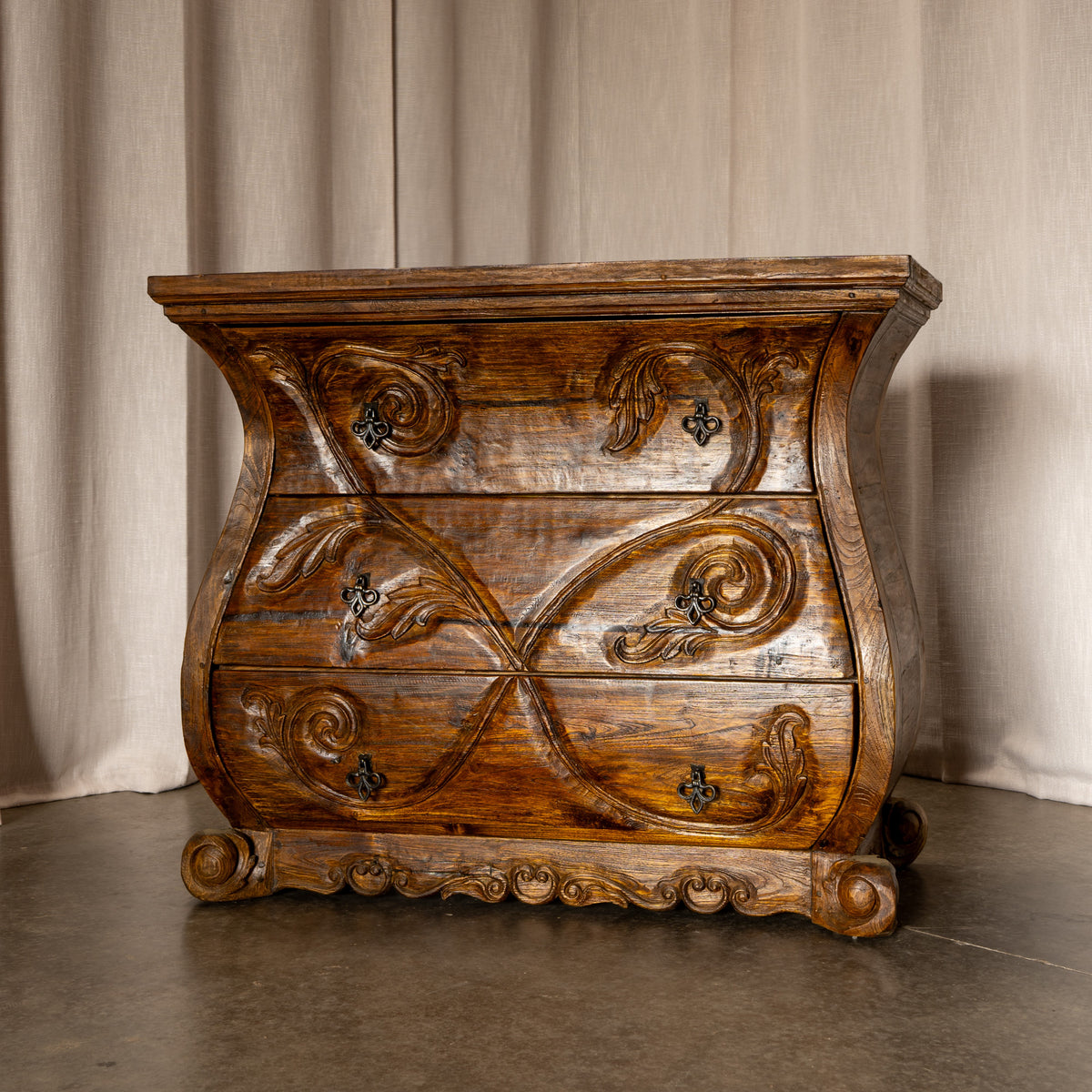 An ornate, curved three-drawer wooden chest of drawers with carved scrollwork and dark metal handles, set against a neutral beige curtain backdrop. Jason Scott Furniture - Angelina Dresser -Fiesta Furnishings. 