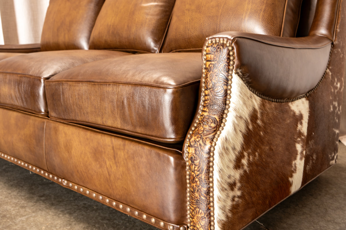 Close-up of a brown leather sofa with carved nailhead trim and a brown-and-white cowhide upholstered side panel, shown indoors on a carpeted floor.