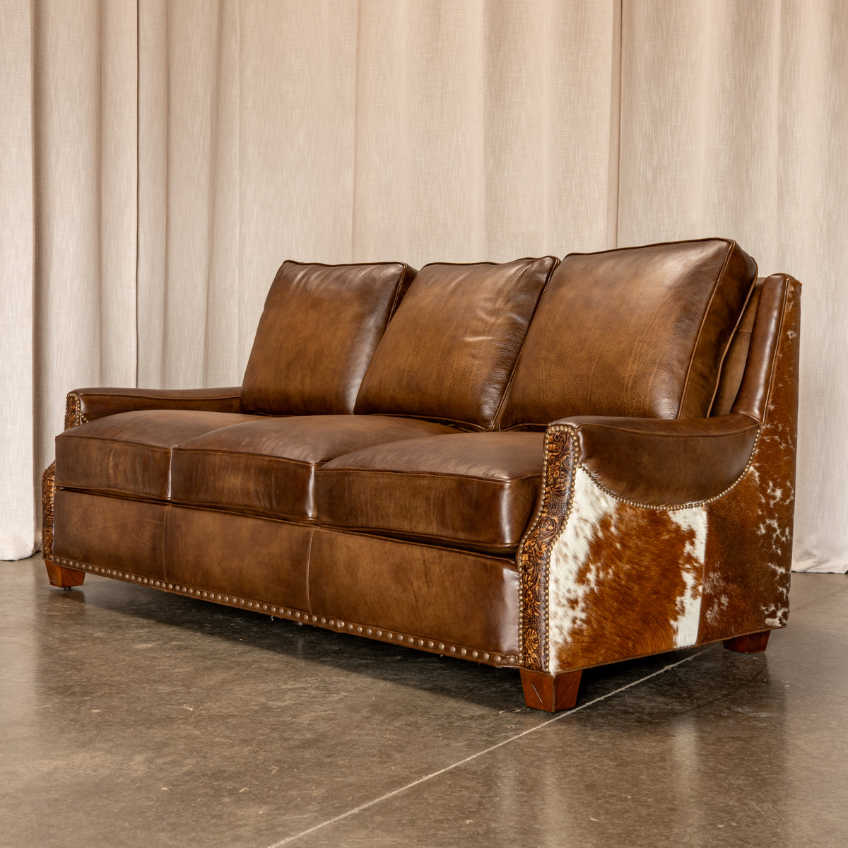 A brown leather loveseat sofa with a cowhide-patterned armrest and nailhead trim, photographed in a studio setting with a beige curtain backdrop and concrete floor.
