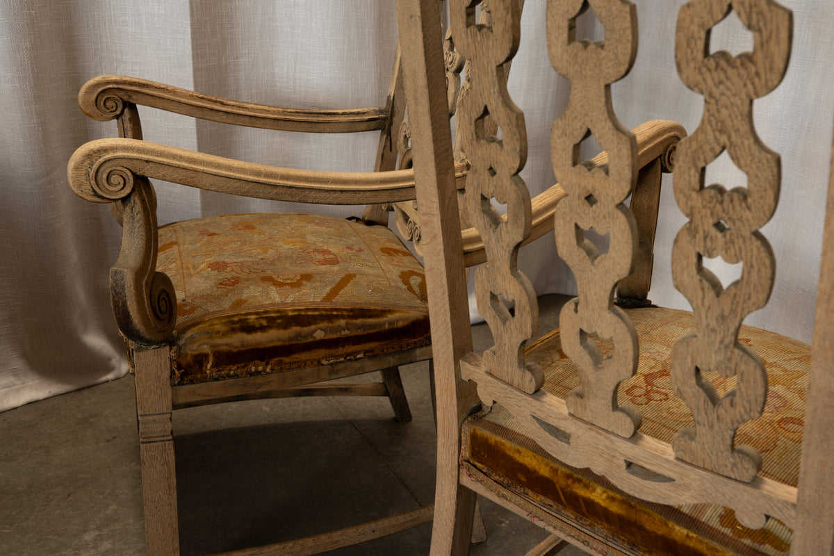 An antique wooden armchair with carved scroll arms and a distressed beige frame, paired with a patterned mustard-yellow and rust upholstered seat cushion, shown next to another matching chair.