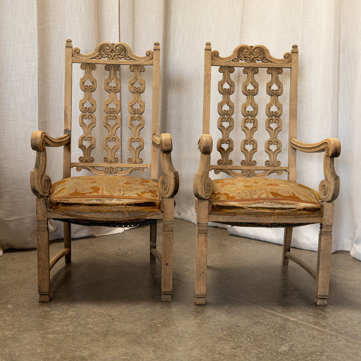 A pair of vintage carved wooden armchairs with ornate scrollwork backrests and distressed tan upholstery, set against a draped beige fabric backdrop.