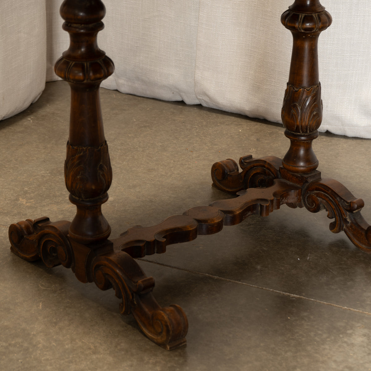 Close-up of an ornate dark brown carved wooden table base with scrollwork and a central stretcher, set on a concrete floor in front of a light fabric backdrop.