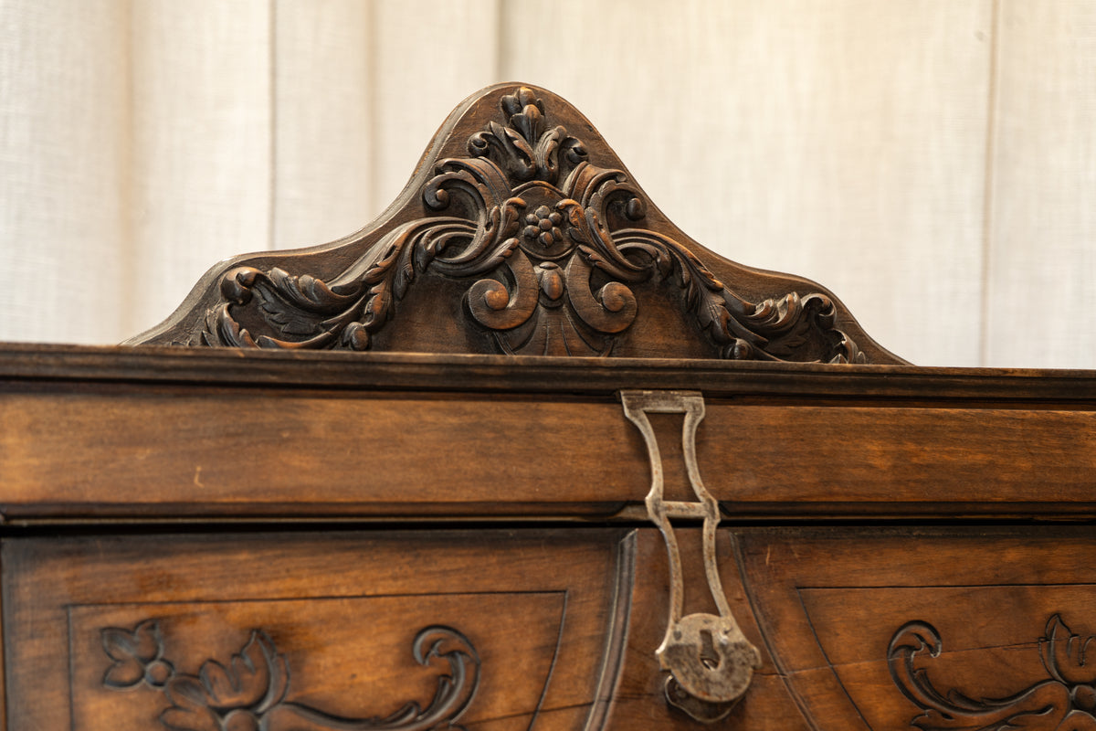Close-up of an ornate dark brown wooden cabinet or hutch, showing carved floral scrollwork on the top panel and a metal latch, against a light beige fabric background.