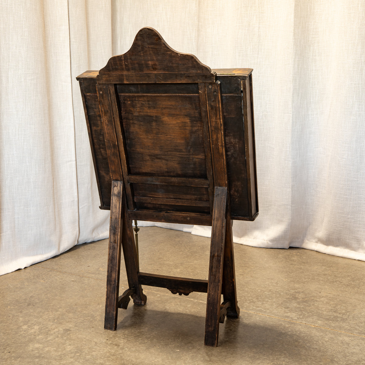 A dark brown wooden folding table or easel with a tall arched backrest, shown from the back against a draped white fabric backdrop and concrete floor.