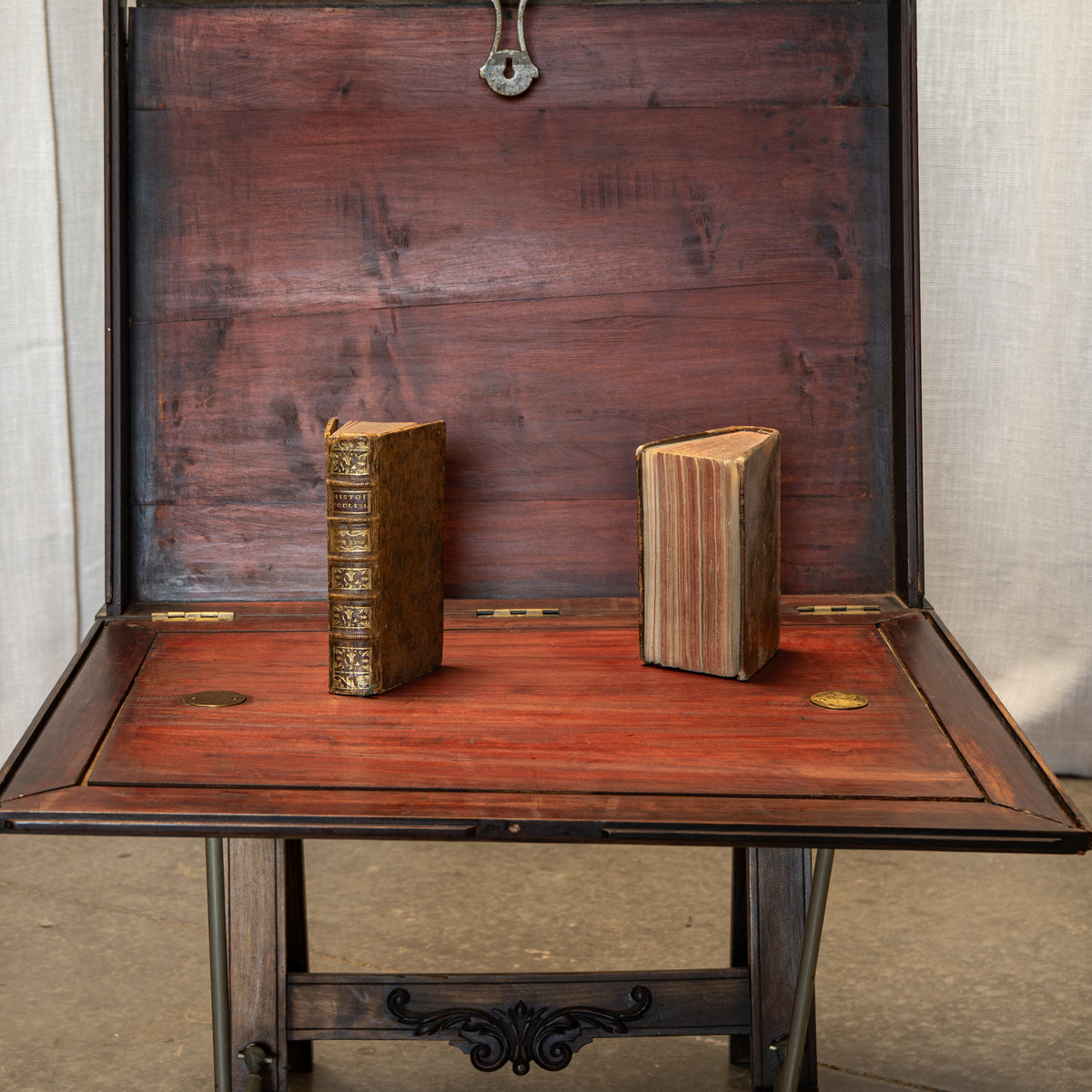 An antique wooden writing desk or hutch with its interior open, showing a reddish-brown wood grain, brass hardware, and two old books standing upright inside.
