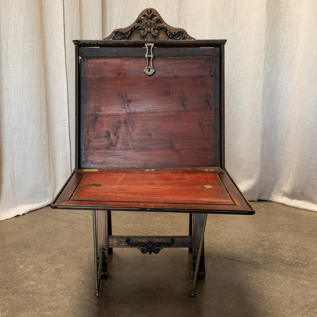 An antique wooden writing desk with an open hinged lid and folded front panel, featuring dark metal legs and brass hardware, set against a neutral draped fabric backdrop.