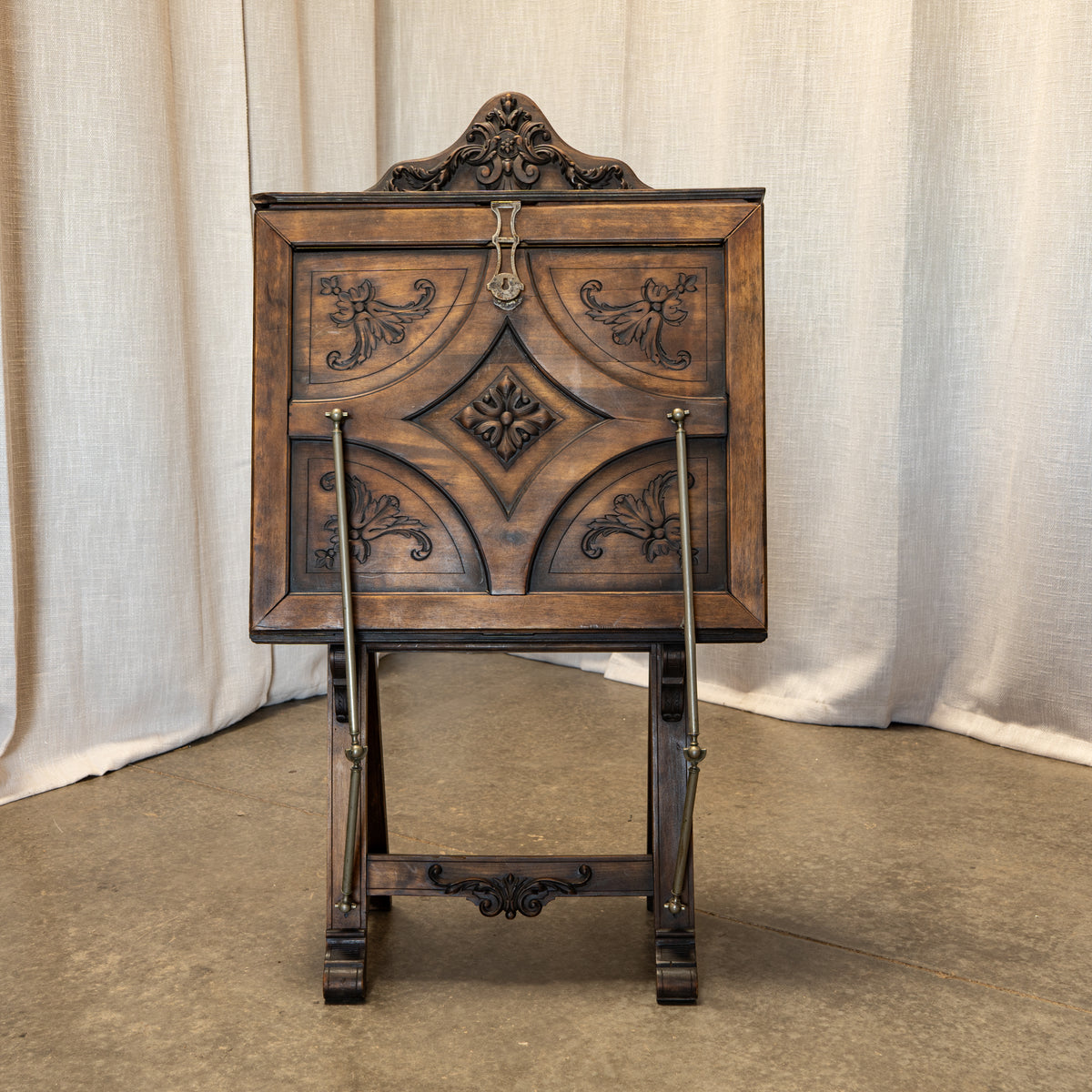 An antique dark brown wooden display stand or easel with ornate carved floral details and metal support rods, photographed against a draped beige fabric backdrop.