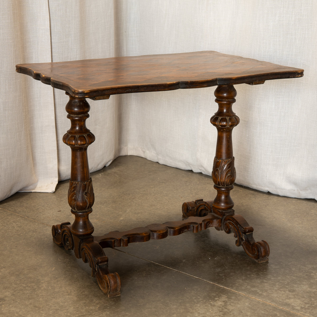 An antique-style dark brown wooden table with carved legs and a scalloped apron, photographed against a draped beige fabric backdrop.