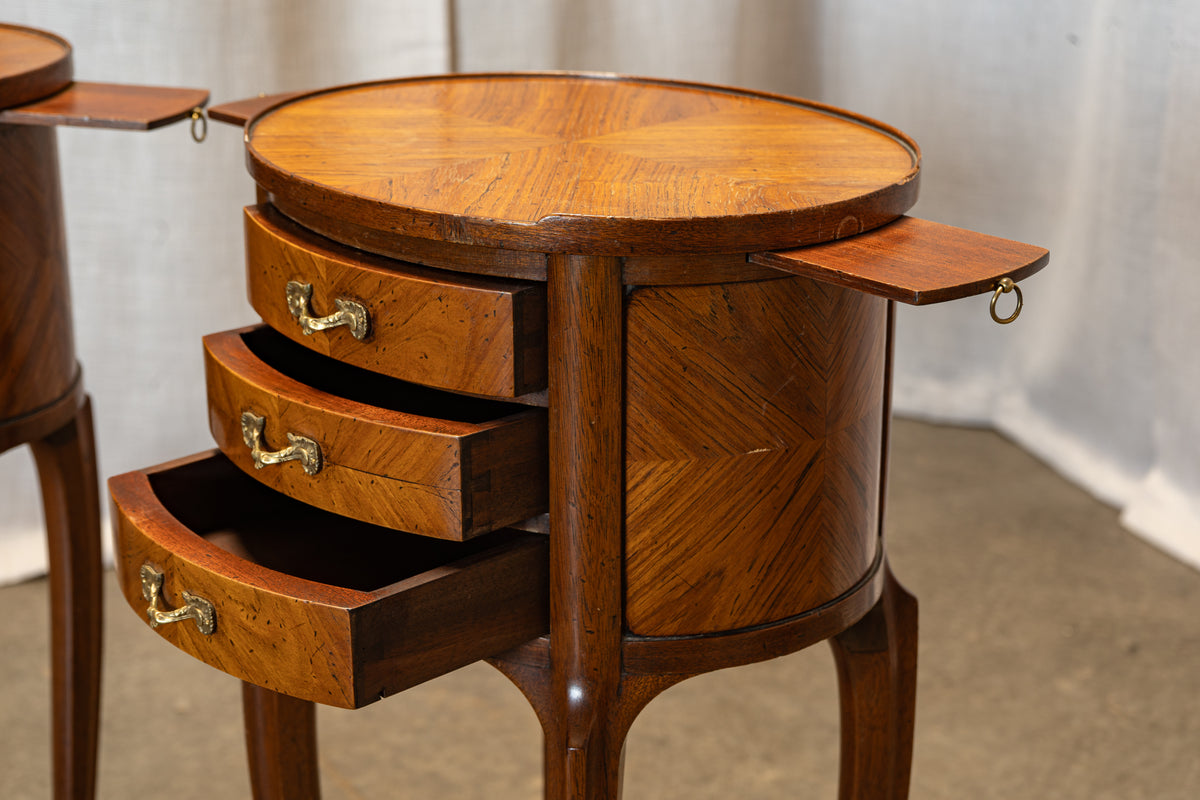 A close-up of a polished wooden side table with three drawers, featuring a round top, inlaid wood patterns, and pull-out side wings with hanging rings, set against a neutral indoor backdrop.