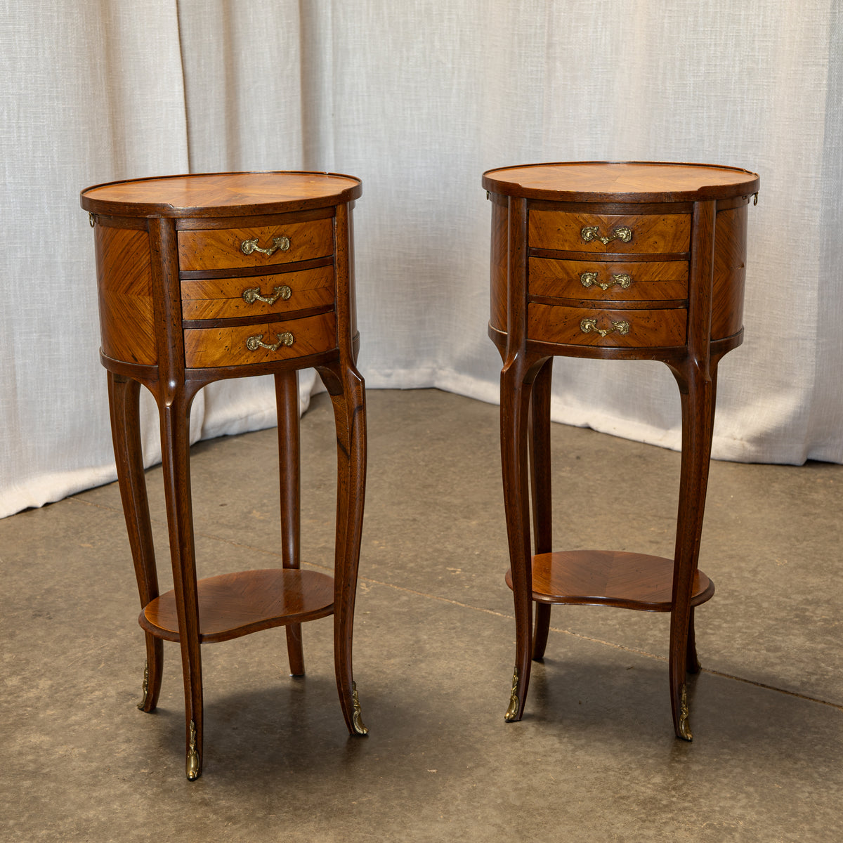 A pair of vintage wooden nightstands with three drawers, curved legs, and brass hardware, set against a neutral draped fabric backdrop.