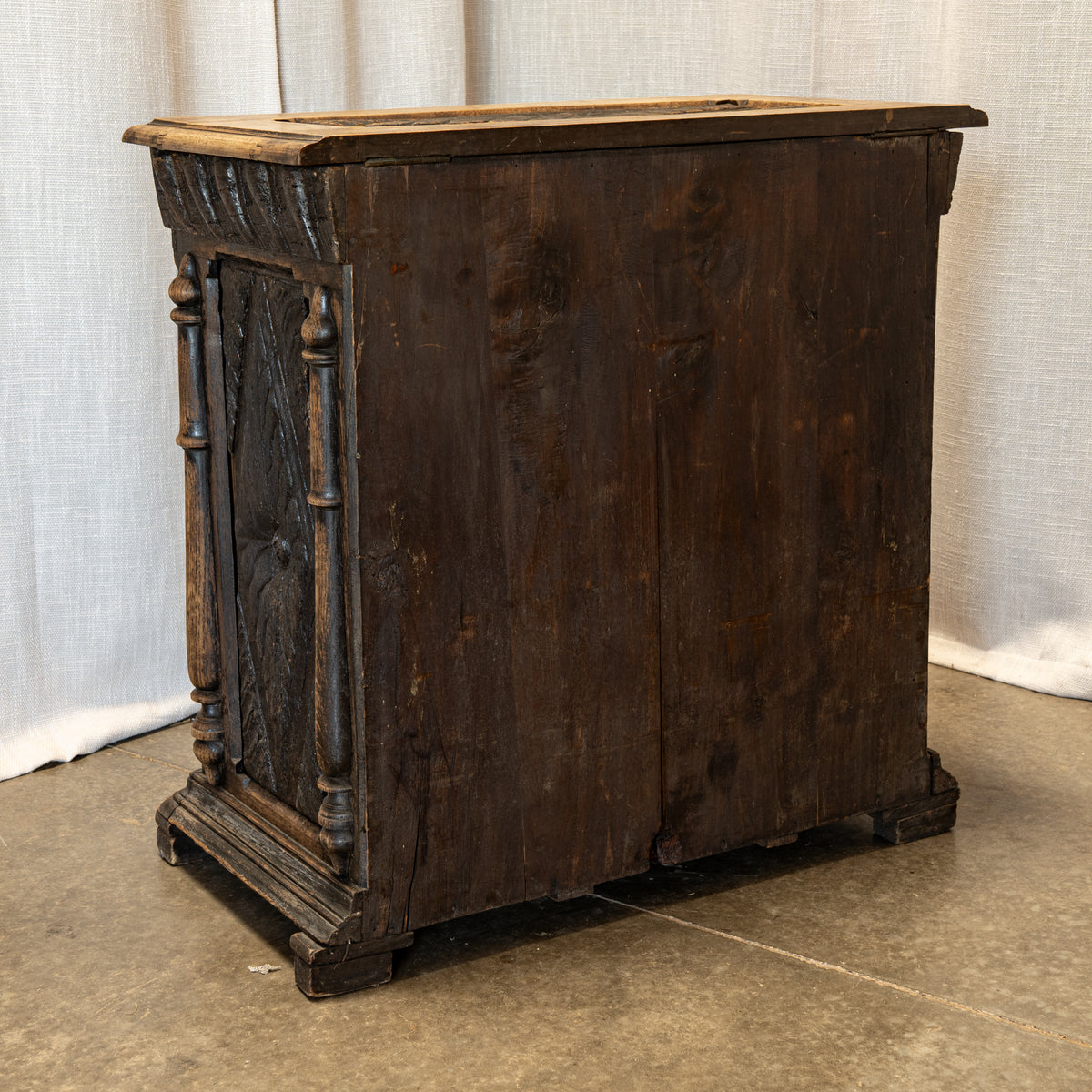 An antique dark brown wooden chest or cabinet with carved decorative side panels and a hinged front, photographed indoors against a light fabric backdrop and concrete floor.