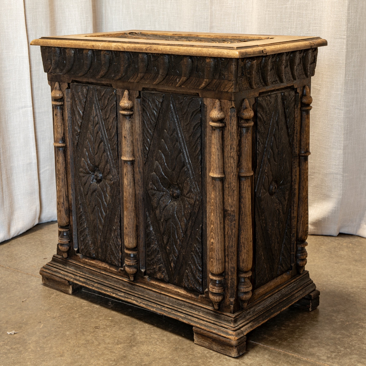 An ornate dark brown carved wooden chest or cabinet with decorative panel doors, turned posts, and a rectangular top, photographed indoors against a light fabric backdrop and concrete floor.