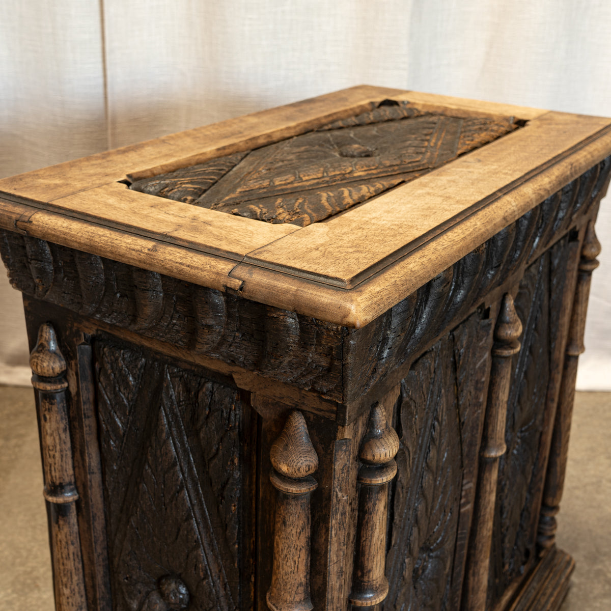 An antique carved wooden chest or cabinet with a dark stained body and a lighter natural wood top, photographed against a neutral draped fabric backdrop.