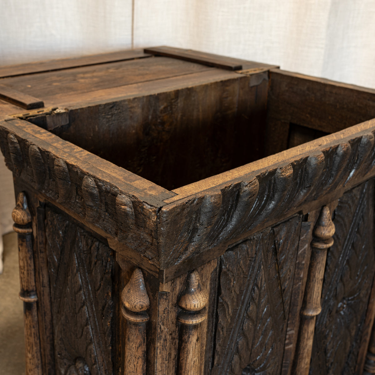 Close-up of a dark carved wooden storage chest or crate with a hinged lid, showing ornate corner posts and a rustic finish against a light fabric backdrop.