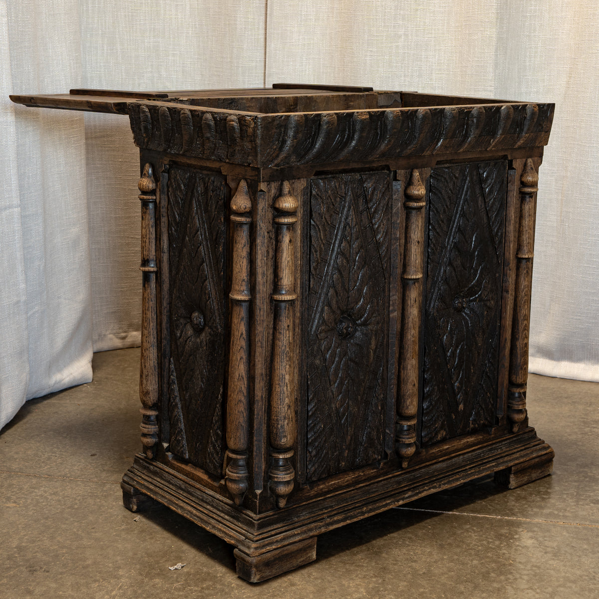 An antique carved wooden chest or cabinet with dark stained panels and turned posts, photographed indoors against a light fabric backdrop and concrete floor.